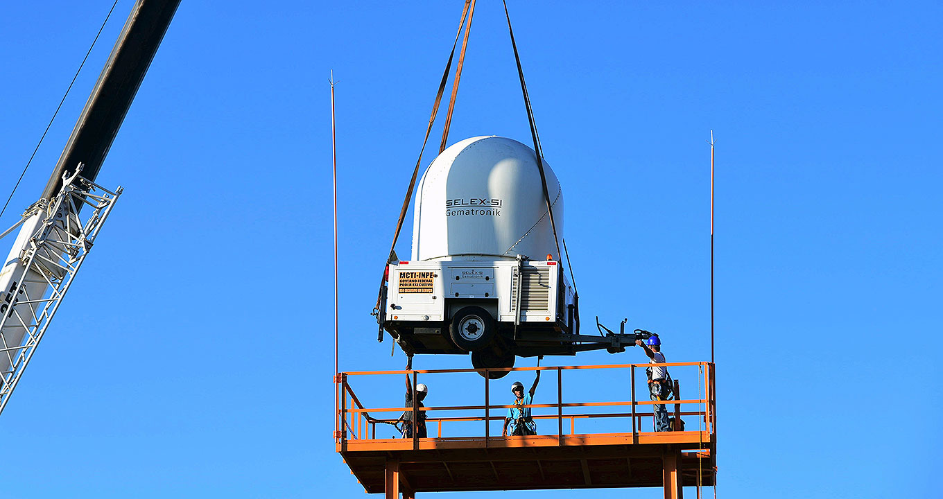 Radar instalado na Unicamp, em outubro de 2016, no âmbito do projeto temático SOS Chuva | Foto: Antonio Scarpinetti Audiodescrição: Em área externa, guindaste com cerca de vinte metros de altura, à esquerda, iça equipamento de formato cilíndrico e arredondado na parte de cima, suspendendo-o com cabos para uma ampla base de ferro, com quatro pés e uma plataforma de ferro. Três pessoas sobre a plataforma de ferro participam da operação. Imagem 1 de 1