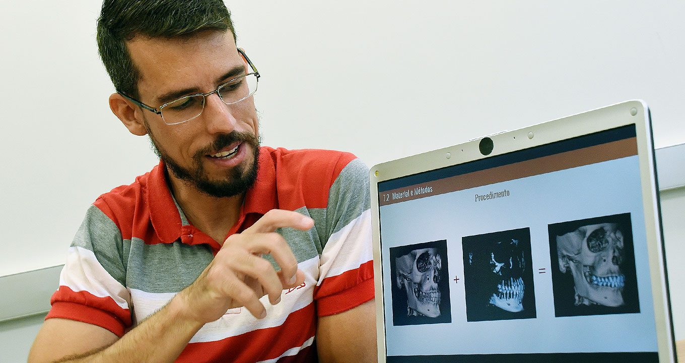 Audiodescrição: Em sala, imagem frontal e de busto, homem sentado, à esquerda na imagem, aponta com o indicador da mão direita para três foto de crânios, dispostas uma ao lado da outra, que são exibidas na tela de um notebook que se encontra à direita na imagem sobre suposta mesa. Ele usa óculos e veste camisa de mangas curtas, com listra horizontais nas cores vermelho, cinza e branco. Imagem 1 de 1.