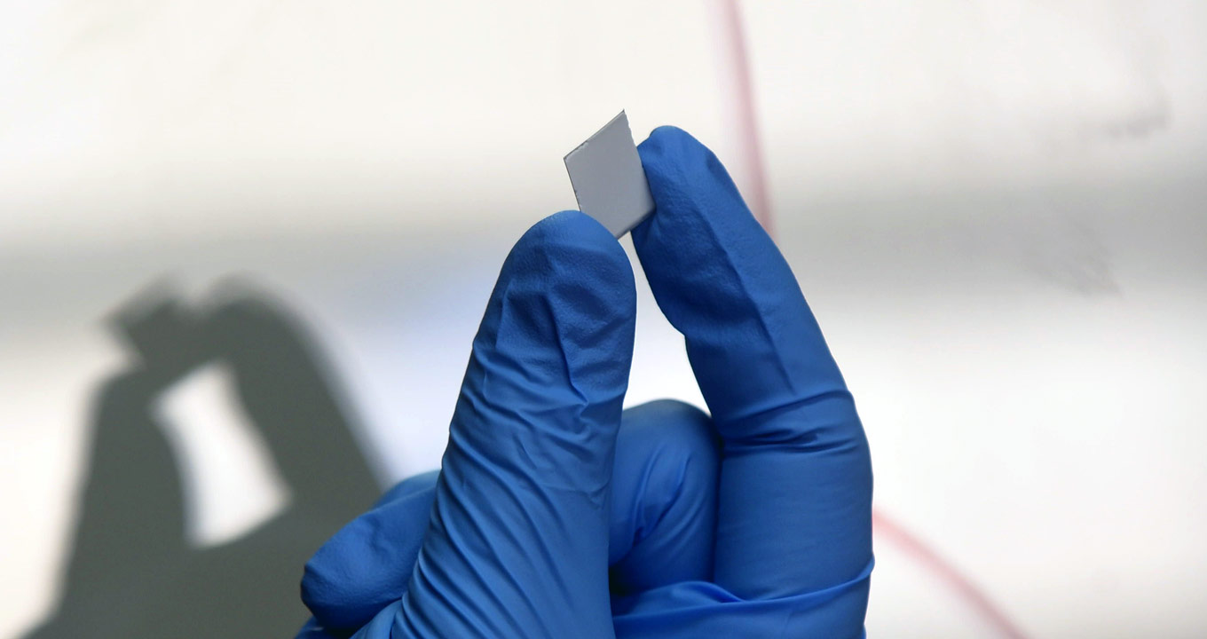 Placa de sílica: selecionando biomarcadores | Foto: Antonio Scarpinetti Audiodescrição: Em área interna, imagem close-up e frontal, dedos da mão direita, utilizando uma luva azul, seguram, com o polegar e o indicador, uma pequena placa de sílica retangular prateada, com cerca de um centímetro por um centímetro. Ao fundo, à esquerda, aparece a sombra dessa mesmo imagem. Imagem 1 de 1.