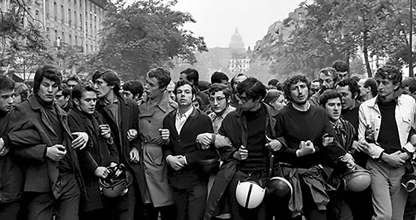 Estudantes marchando em Paris", em 1968 | Foto: Henri Cartier Bresson | Reprodução Editora Unesp Audiodescrição: Em imagem preto e branco, frontal e em plano americano, cerca de cinquenta pessoas, em pé, uma encostada à outra, de braços dados, agrupadas horizontalmente em uma grande manifestação. Todos usam roupas de frio, como blazers, jaquetas e sobretudos, e alguns levam um tipo de capacete. Ao fundo, ao centro, há uma torre de uma edificação clássica, à esquerda uma edificação, e à direita um canteiro com árvores altas. Imagem 1 de 1.