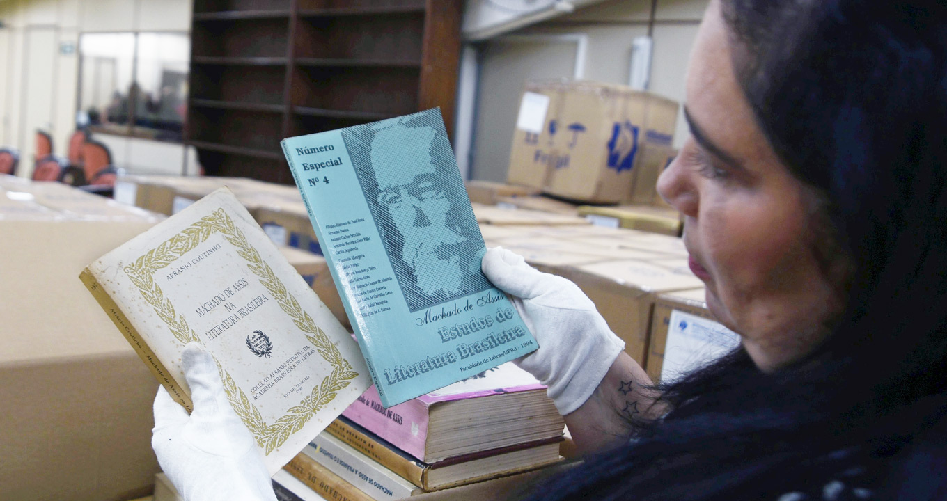 A bibliotecária Maria Helena Segnorelli confere conteúdo de caixa com livros da biblioteca de Antonio Candido | Foto: Antonio Scarpinetti Audiodescrição: Em ampla sala, imagem close-up, em perspectiva e de trás para frente, mulher, à direita na imagem, segura com as mãos dois livros de cerca de vinte centímetros de altura, por quinze de largura, com as capas voltadas para frente. Nelas se lê Número especial quatro, Machado de Assis, e Machado de Assis na Literatura Brasileira. Ela usa luvas brancas e mantém os braços erguidos na altura do rosto. À frente dela, ocupando quase toda sala, há várias caixas de papelão empilhadas. Imagem 1 de 1.