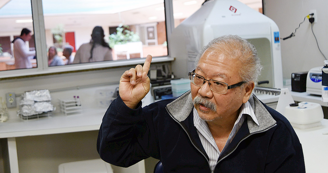 Audiodescrição: Em laboratório médico, imagem frontal e de busto, homem sentado, ao centro na imagem, fala gesticulando com o braço direito, mantendo-o recolhido e levemente erguido para o alto, apontando com o dedo indicador para cima. Às costas dele, na junção das paredes, há uma bancada de madeira na cor branca, sendo que sobre ela há vários equipamentos de laboratório. Ao fundo, à esquerda na imagem, há uma janela de vidro transparente, permitindo ver outras pessoas em pé do lado de fora. Imagem 1 de 1.