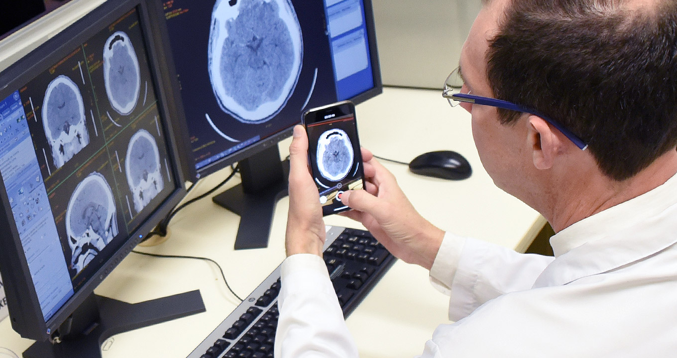 O neurocirurgião Luiz Adriano Esteves, autor da pesquisa | Foto: Antoninho Perri Audiodescrição: Em laboratório médico, imagem em perspectiva, de costas e em close-up, de homem sentado em cadeira, à direita na imagem, segura smartphone com as duas mãos, fotografando imagem de um crânio, reproduzida em exame, e aparece em monitores dispostos à frente dele, sobre uma mesa de madeira. Sobre a mesa estão os dois monitores de lcd, dispostos um ao lado do outro, teclado e mouse. Ele usa óculos e veste jaleco branco de mangas compridas. Imagem 1 de 1.