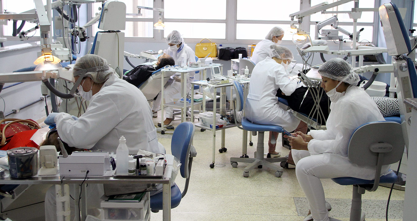 Ambulatório da Faculdade de Odontologia de Piracicaba | Foto: Antonio Scarpinetti Audiodescrição: Em amplo consultório odontológico, imagem em perspectiva e de corpo inteiro, quatro dentistas prestam atendimento odontológico a pacientes que estão sentados em cadeiras de dentista. Os dentistas estão dispostos um à frente do outro, sendo dois à esquerda e dois à direita. Todos usam jalecos brancos de mangas compridas, toucas, luvas e máscaras. Ao fundo uma ampla janela horizontal de vidro transparente e estrutura metálica branca, formada por módulos horizontais. Imagem 1 de 1.