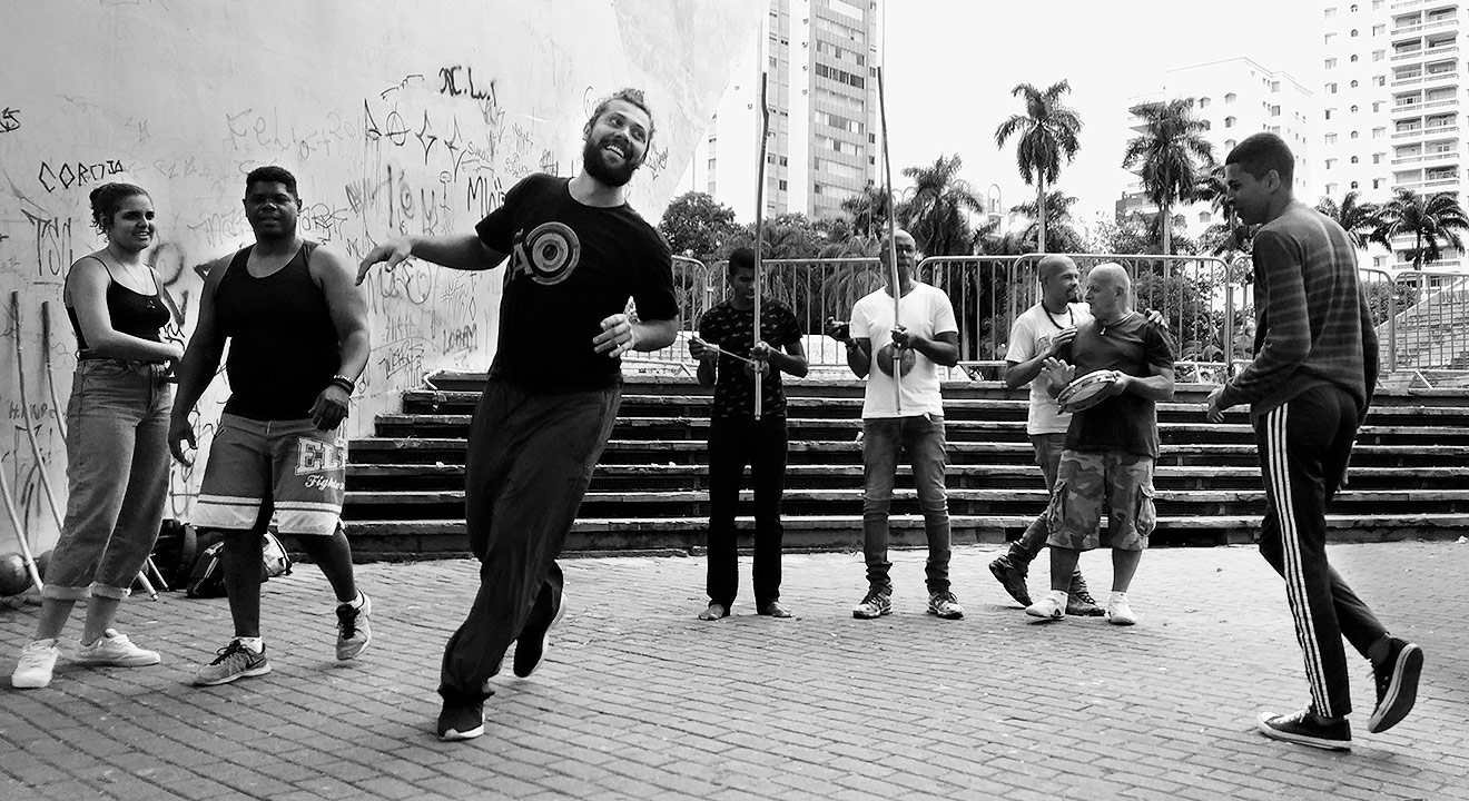 Roda de capoeira no Centro de Convivência Audiodescrição: foto preto e branco, área externa, imagem de corpo inteiro e frontal, oito pessoas em pé, próximas uma da outra, sendo que três delas, ao fundo, tocam instrumentos, sendo dois berimbaus e um pandeiro. À frente deles, dois homens parecem dançar, enquanto outros os observam. À esquerda na imagem, há um grande e alto paredão, e ás costas dessas pessoas, unida ao muro, extensa escadaria com sete degraus, cerca de dez grades de proteção móveis, de ferro, altas palmeiras e prédios. Imagem 1 de 1.