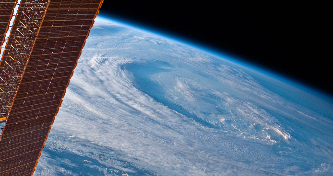 Imagem do espaço, onde se vê parte do planeta Terra, com superfície repleta de nuvens e massas de ar em espiral, tendo ao fundo o espaço, em preto. À esquerda na imagem, parte de um satélite. Imagem 1 de 1.