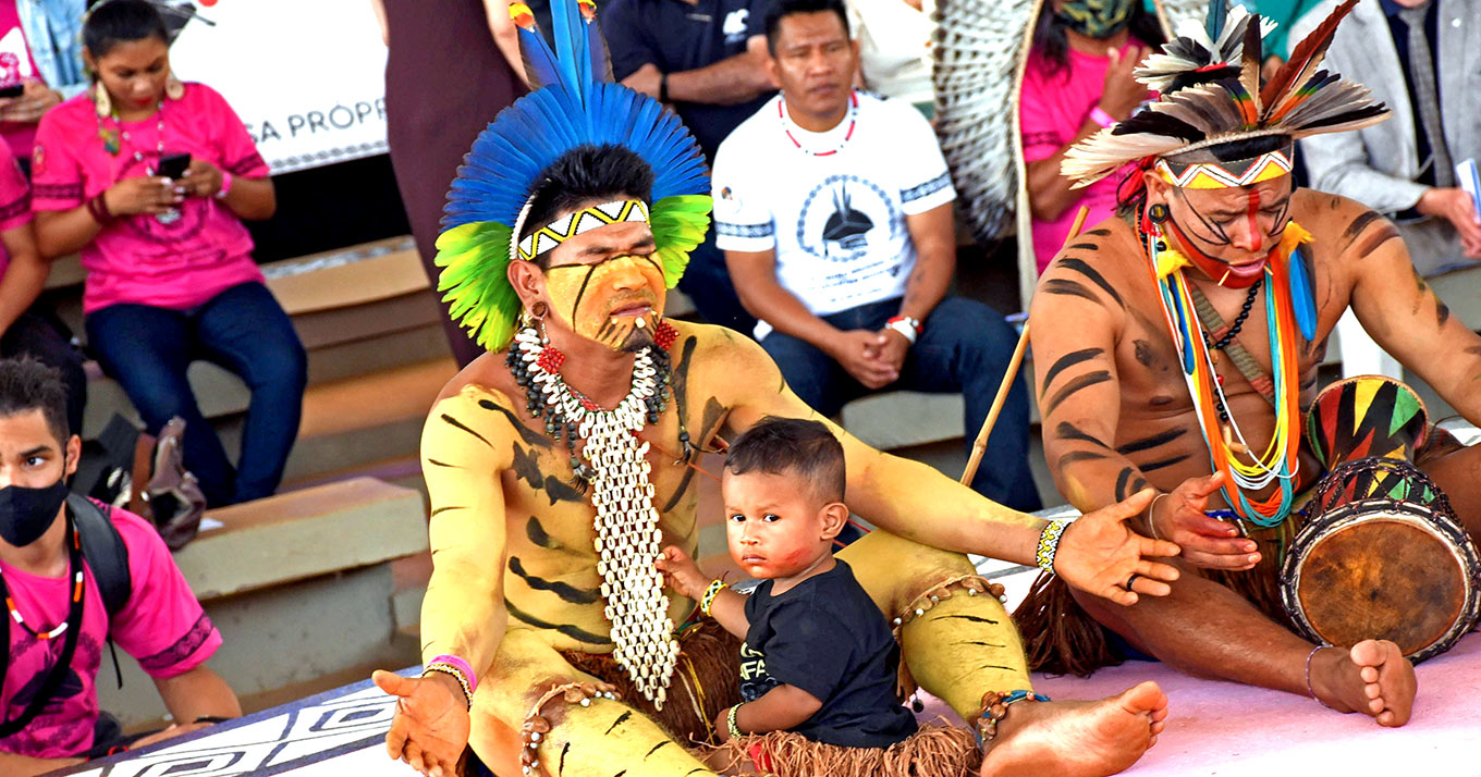 Foto com dois homens indígenas que estão sentados, com as pernas cruzadas e de olhos fechados. Na frente do homem à esquerda há uma criança sentada.