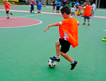 Foto de um grupo de crianças em um treino de futubol. No primeiro plano aparece um menino branco que usa shorts e colete laranja.  Há uma bola de futebol sob os pés dele.