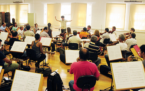 músicos da orquestra ensaiam em sala. Maestro ao fundo rege.