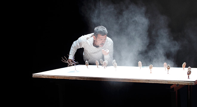 Imagem de espetáculo do artista Gustavo Ciríaco. Sobre uma mesa ele sopra uma nuvem de poeira.