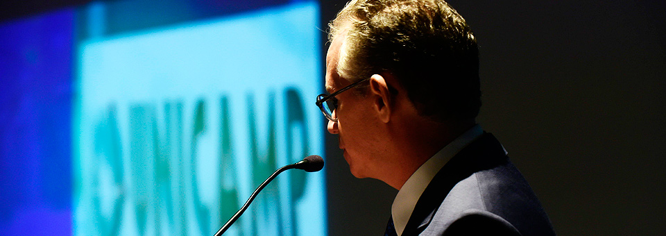 O reitor Marcelo Knobel em discurso de posse De perfil ao centro da foto o Professor Marcelo Knobel em discurso próximo ao microfone, ao fundo imagem do logo e o nome da Unicamp projetada na tela , cor de fonte preta e fundo branco