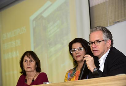 O reitor, Marcelo Knobel; a coordenadora do Cocen (Coordenação de Centros e Núcleos), Ana Carolina de Moura; e a professora Graça Caldas, na mesa de abertura do lançamento do livro Em auditório, em perspectiva, três pessoas sentadas em cadeiras, uma ao lado da outra, sendo que o homem, à direita, fala em microfone sem fio, segurando-o com a mão esquerda. À frente deles, há uma mesa retangular de madeira, e às costas, à direita, uma grande tela de projeção de data show que exibe a capa de um livro.