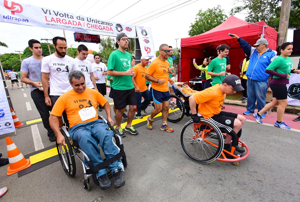 Imagem com a largada dos corredores cadeirantes da Volta da Unicamp de 2015. Na foto, três cadeirantes estão junto com outros atletas, de ambos os sexos, passando pela largada sentido direita.