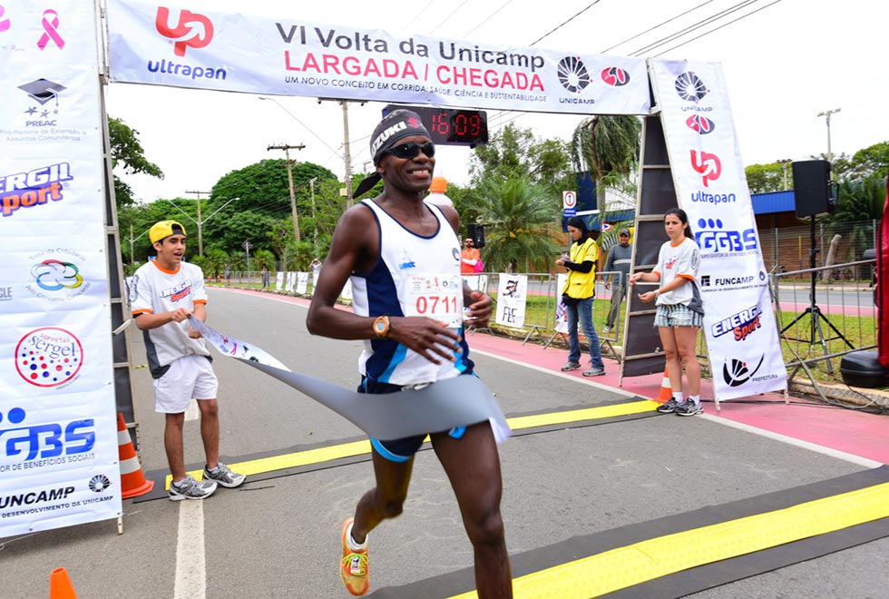 Imagem de um corredor homem cruzando a linha de chegada da edição de 2015 da Volta da Unicamp. Na foto ele corre para a direita enquanto passa pela faixa que sinaliza o fim da corrida segurada por membros da organização da prova 