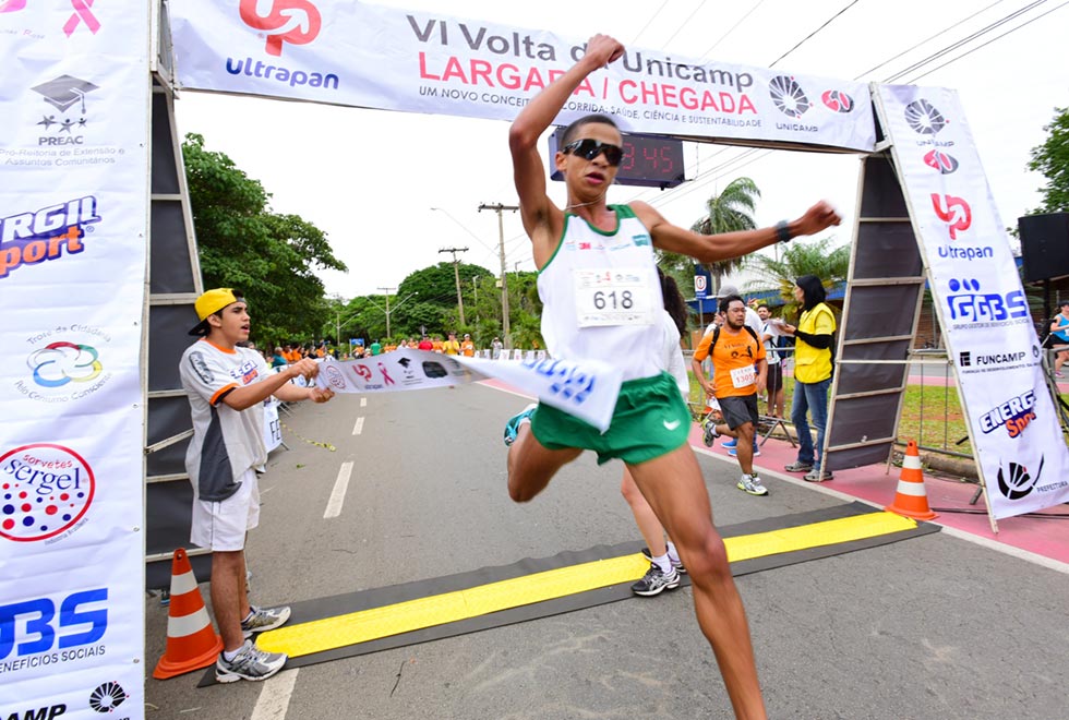 Imagem de um corredor homem cruzando a linha de chegada da edição de 2015 da Volta da Unicamp. Na foto ele pula em direção para a direita com os braços levantados enquanto passa pela faixa que sinaliza o fim da prova segurada por membros da organização