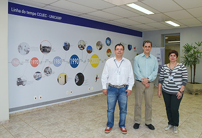 Sandro Rigo, Rubens Queiroz de Almeida e Maria Inês Santi´Anna frente à linha do tempo da história do CCUEC dois homens e uma mulher em pé frente a um painel com uma linha do tempo