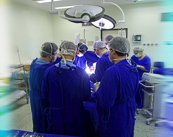 Equipe do centro cirúrgico do Hospital de Clínicas da Unicamp realiza cirurgia. Na foto, nove profissionais estão paramentados com jalecos azuis e toucas brancas em volta de um paciente que está sendo operado na mesa.