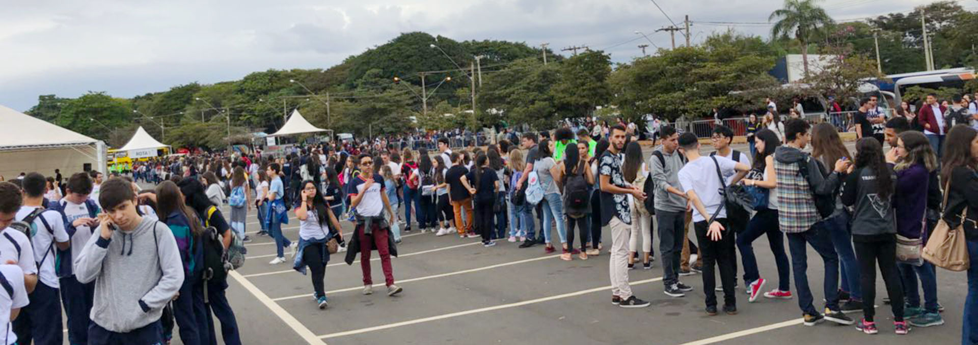 Audiodescrição: Em área externa, imagem panorâmica e em perspectiva, extensa fila, ao centro, com centenas de jovens em pé, conversando, sendo que há outras filas paralelas a essa. Eles estão em uma ampla área de estacionamento com pintura de demarcação das vagas no chão. As filas seguem da direita para a esquerda. Há três tendas de lona branca à esquerda, e ao fundo de toda a imagem uma densa área verde, com árvores de grande porte. O tempo está nublado. Imagem 1 de 1.