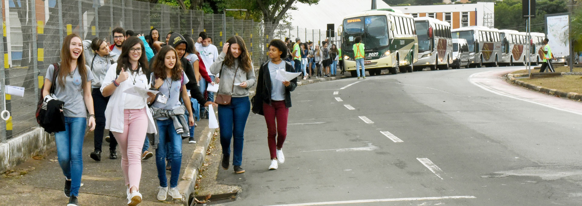 Audiodescrição: Em área externa, imagem panorâmica e frontal, cerca de quarenta jovens, à esquerda na imagem, caminham em calçada de cimento, junto à rua asfaltada, em trecho de curva. Eles desembarcaram de veículos, cinco ônibus e uma van, que estão estacionados na rua, ao fundo na imagem. Há uma grade de ferro que contorna toda a calçada. Os jovens em primeiro plano na fila sorriem. Imagem 1 de 1.