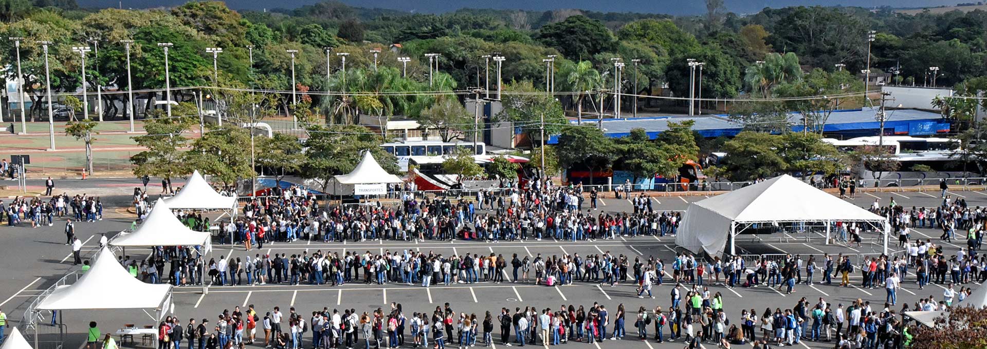 Estudantes chegam para UPA Audiodescrição: Em área externa, imagem panorâmica do alto para baixo, pátio de estacionamento de veículos asfaltado e com pintura de demarcação das vagas, onde estão centenas de pessoas, em pé, formando três grandes filas que se estendem da esquerda para a direita. À frente de cada fila há uma tenda com lona branca. Ao fundo, paralelos às filas, há vários ônibus estacionados um à frente do outro e também vários refletores instalados em postes, e uma ampla área de vegetação com altas árvores. Imagem 1 de 1.