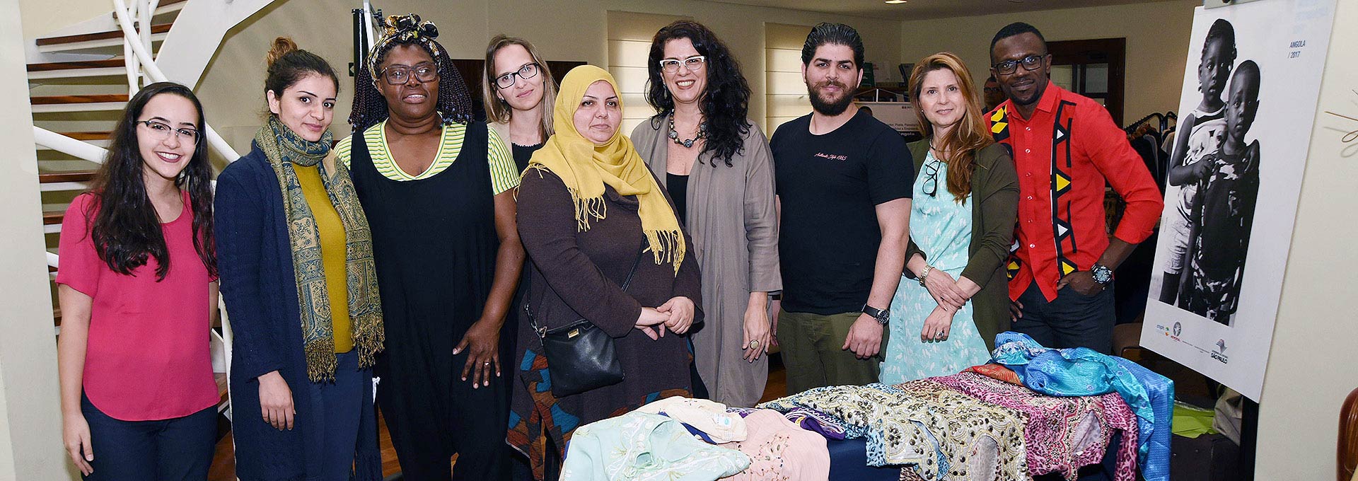Organizadores do evento e participantes da feira de produtos dos refugiados | Foto: Antonio Scarpinetti Audiodescrição: Em área interna, imagem frontal e em plano médio, nove pessoas em pé, uma ao lado da outra, sendo que quase todos sorriem com olhares voltados para a câmara fotográfica. À frente delas, há uma bancada retangular com vários roupas e tecidos estampados coloridos. Alguns vestem trajes típicos do oriente médio e áfrica. À direita na imagem, aparece, afixado na parede, parte um banner vertical com uma foto preto e branco de duas crianças. Imagem 1 de 1.