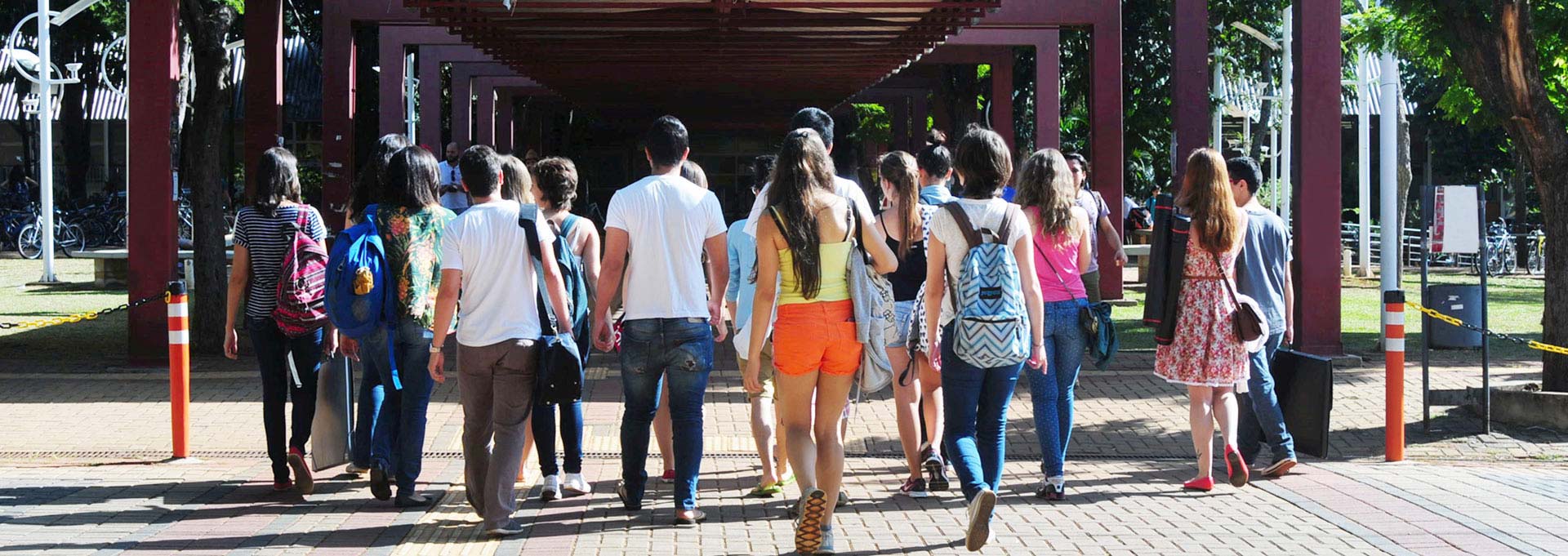 Mais estudantes passaram a receber bolsas este ano Audiodescrição: Em área externa ensolarada, imagem de corpo inteiro, grupo de cerca de vinte jovens, de costas na imagem, caminha para frente sobre amplo calçamento de piso intertravado de concreto. A maioria usa camiseta, short ou calça jeans, e carrega mochila nas costas. À frente deles há um corredor formado por estruturas metálicas semelhantes a um gol de futebol, com duas traves e um travessão, com cerca de dez metros de largura por quatro de altura, dispostas uma à frente da outra. Imagem 1 de 1.