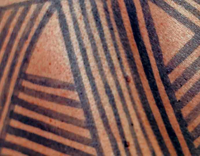 Audiodescrição: Em imagem close-up e frontal, pintura indígena brasileira feita na pele de uma pessoa com uso de tinta preta e elaborada com traços verticais e na diagonal, unidos, como que formando triângulos. Imagem 1 de 1.