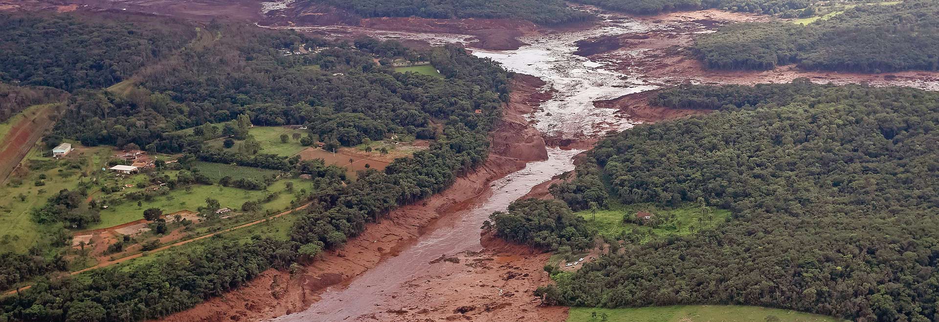 Região atingida pelo rompimento da barragem Mina Córrego do Feijão, em Brumadinho/MG. Publicado em 26/01/2019 - 12:07 | Foto: Agência Brasil - Divulgação Audiodescrição: Em imagem panorâmica, foto aérea de amplas áreas com vegetação à esquerda e à direita, sendo que ao centro há um rio repleto de lama que cruza a imagem, vindo de cima para baixo. Há uma grande área com lama e barro na parte de cima de imagem. Imagem 1 de 1.