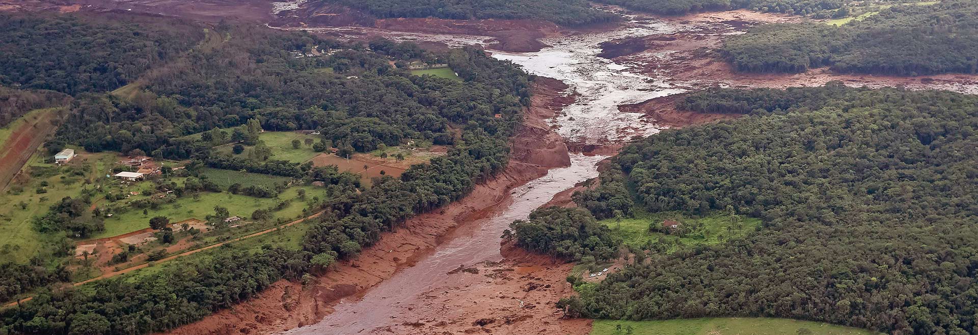 Audiodescrição: Em imagem panorâmica, foto aérea de amplas áreas com vegetação à esquerda e à direita, sendo que ao centro há um rio repleto de lama que cruza a imagem, vindo de cima para baixo. Há uma grande área com lama e barro na parte de cima de imagem. Imagem 1 de 1.