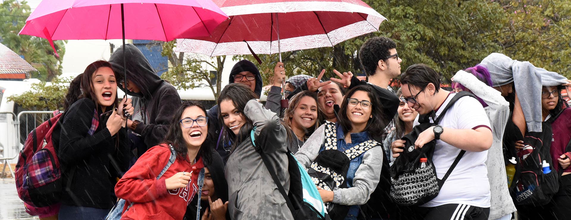 Audiodescrição: Em área externa, imagem frontal e em plano médio, grupo de cerca de quinze jovens, em pé, agrupados horizontalmente, sendo que a maioria sorri e carrega mochilas nas costas e à frente, e também veste blusas de mangas compridas com capuz. Chove forte, e eles se abrigam sob dois guarda-chuvas, tentando se proteger. Ao fundo, À direita, há uma ampla área com vegetação, e à esquerda, parte de uma edificação. Imagem 1 de 1.