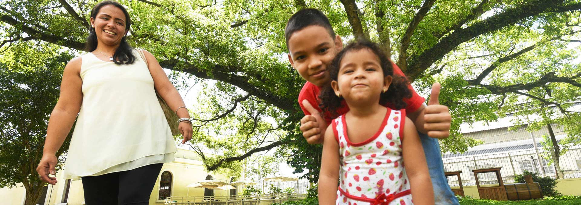 Audiodescrição: Em área externa, imagem frontal de baixo para cima e em plano médio, duas crianças, ao centro na imagem, sendo um menino que encontra-se inclinado para frente e faz sinal de positivo com as duas mãos, tendo logo à frente, entre os braços, uma menina com cerca de três anos de idade, em pé, que sorri. Ambos são observados por uma mulher, à esquerda, que está em pé e sorri. Logo atrás deles, em todo o fundo da imagem, uma árvore de grande porte, com muita folhagem e longos galhos. Imagem 1 de 1