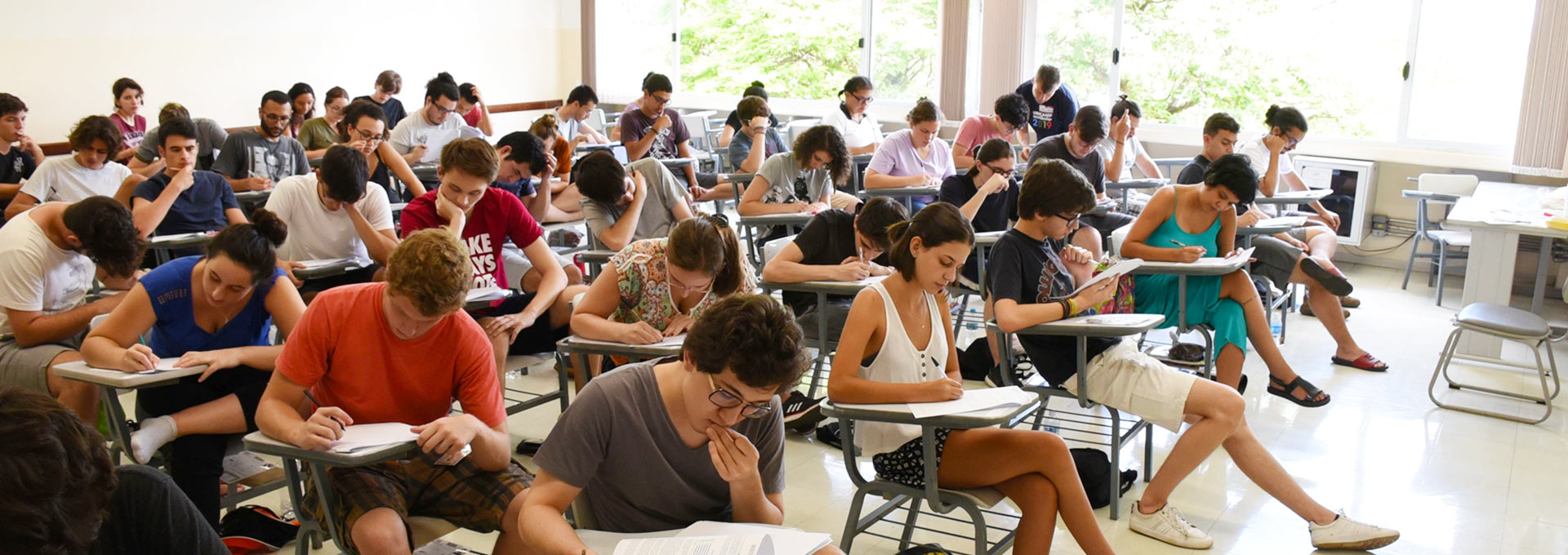 Audiodescrição: em ampla sala de aula, imagem de corpo inteiro e em perspectiva, cerca de quarenta jovens, com olhares voltados para a direita, sentados em carteiras escolares brancas e com braço, dispostas uma ao lado da outra, formando fileiras horizontais. Eles fazem provam, sendo que a maioria mantém a cabeça baixada e observam ou escrevem com caneta em folhas de papel que estão sobre as carteiras. Ao fundo, em toda extensão da sala, há uma ampla janela, que permite ver vegetação externa. Imagem 1 de 1.