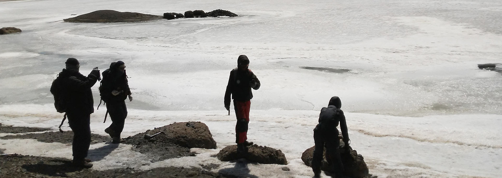 Equipe explorando a ilha em temperatura mais amena Audiodescrição: em área externa no continente antártico, imagem de corpo inteiro e em perspectiva, grupo de quatro pessoas, sendo três em pé, à esquerda e a centro, e uma agachada, à direita, observa a área ao redor, que encontra-se completamente tomada por gelo. Eles estão sobre rochas encravadas na neve e usam roupas especiais para o frio, com capuz. Dois deles usam mochilas às costas, e um deles fotografa o local. Imagem 1 de 1.