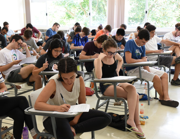 Audiodescrição: Em sala de aula, com janelas ao fundo, grupo estudantes sentados durante prova do vestibular. Tecle enter para acessar