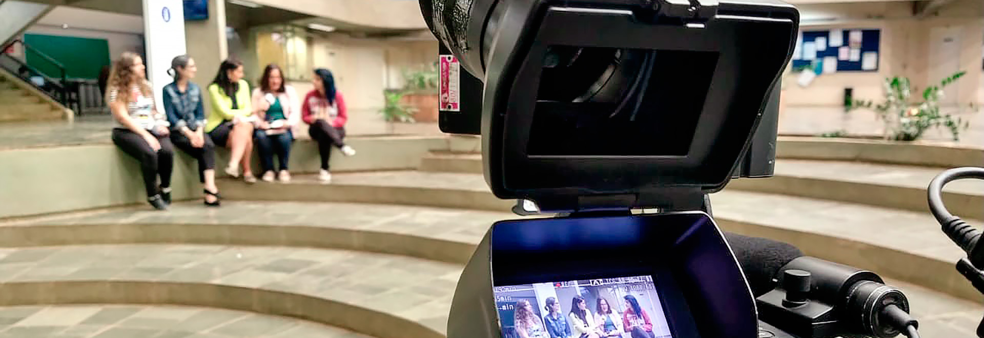 A proposta tem o objetivo de incentivar a carreira científica, especialmente em áreas de exatas e engenharias Audiodescrição: em amplo hall com iluminação natural, imagem frontal e panorâmica, cinco mulheres sentadas em uma mureta de concreto, uma ao lado da outra, em imagem desfoca, à esquerda. A mulher ao centro segura um microfone, entrevistando a mulher à direita. Em primeiro plano, imagem close-up de parte dos visores de uma câmara de filmagem profissional, à direita, que reproduz a imagem das mulheres. O local é edificado com estruturas de concreto, como ampla escada circular, colunas e vigas. Imagem 1 de 1.
