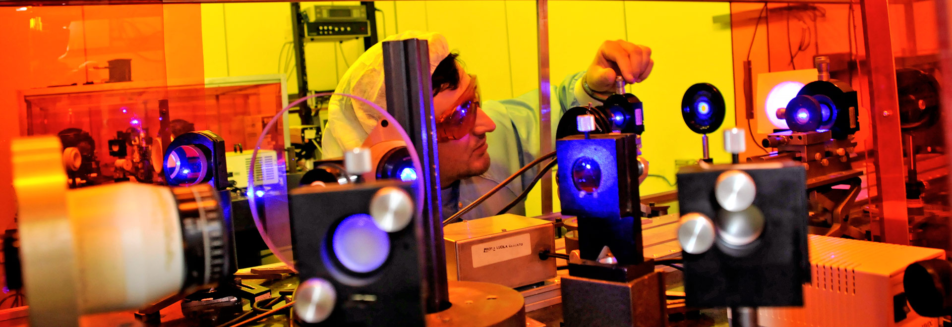 Laboratório de ótica - IFGW | Foto: Antoninho Perri Audiodescrição: em laboratório, imagem de perfil e de busto, homem ao centro da imagem opera com a mão esquerda um equipamento, girando com os dedos indicador e polegar um pino acoplado a um tipo de lente de vidro, com luz no centro. Há vários outros tipos de lentes alinhadas uma à frente da outra, com distância de cerca de 30 centímetros entre elas, sendo que estão sobre uma extensa bancada. A imagem é bem colorida, com luzes laranjas e amarelas. O homem usa luva e roupa especial de proteção. Imagem 1 de 1