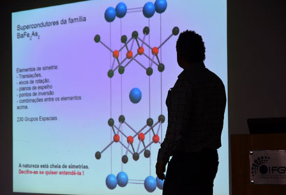 Física para Curiosos atrai público para um bate-papo sobre os átomos Imagem mostra silhueta do professor em foto feita contraluz. Atrás aparece o telão com a imagem dos átomos