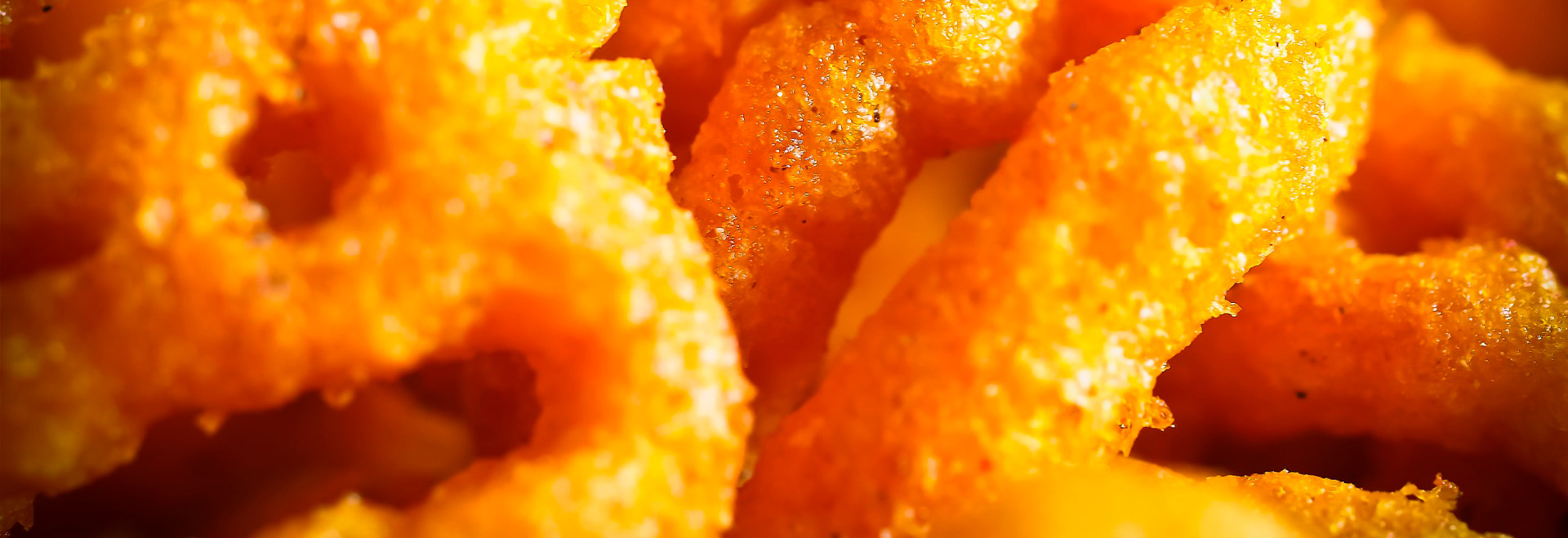 Audiodescrição: em imagem close-up, alimentos fritos em tons amarelados e alaranjados. A imagem é muito aproximada, dificultando identificar qual é o alimento, mas que se assemelha a algum tipo de salgadinho. Imagem 1 de 1.