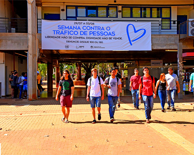 Estudantes caminham no ciclo básico onde vemos, atrás deles, uma grande faixa branca com os dizeres da campanha escritos em azul