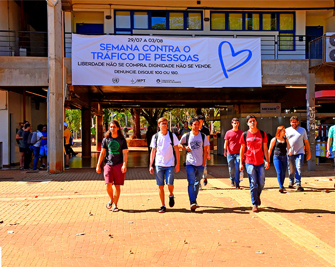 Faixa com dizeres da campanha foi colocada no Ciclo Básico Estudantes caminham no ciclo básico onde vemos, atrás deles, uma grande faixa branca com os dizeres da campanha escritos em azul