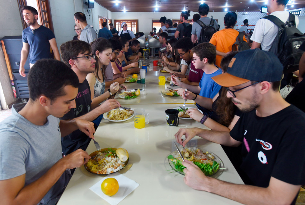 No interior do Restaurante Administrativo da Unicamp, jovens almoçam em uma grande mesa. Quase todos estão utilizando canecas para tomar o suco. 