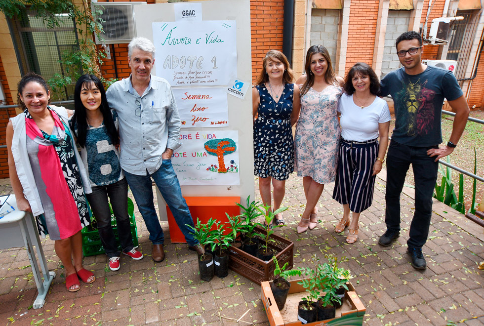 Grupo composto de sete funcionários (cinco mulheres e dois homens) posa para a foto ao lado do cartaz "adote uma árvore". Eles organizaram a ação que distribuiu 50 mudas de plantas.