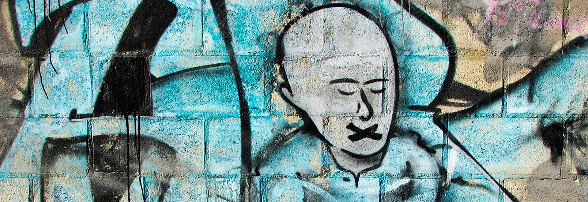 Imagem: Dimitris Vetsikas , domínio público Audiodescrição: em área externa, imagem frontal de um muro construído com blocos de concreto onde há aplicado um grafite colorido. A imagem de grafite traz um homem calvo, com olhos fechados e voltados para baixo, e com um xis na boca. Ele parece estar lendo um livro, o qual segura com as duas mãos. A pintura do corpo do homem é em branco, e a do entorno dele nas cores azul e preto. Imagem 1 de 1.