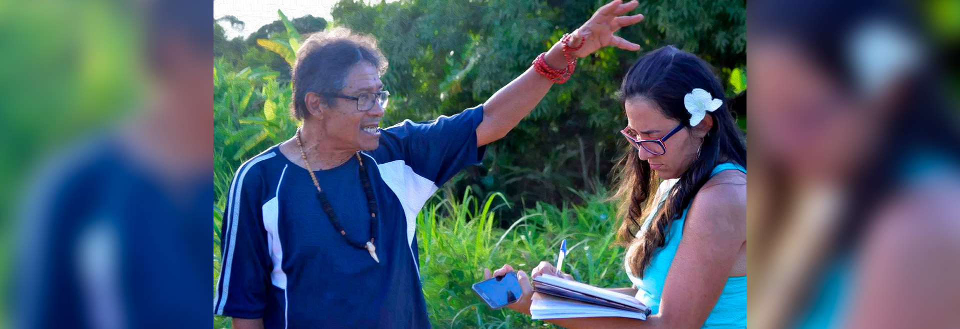 O cacique Juvenal Payayá e Jamille. Memória oral que colaborou para busca de documentos e manuscritos Audiodescrição: em área de mata, imagem em perspectiva e plano médio, homem em pé, com olhar voltado para a direita da imagem, mantém o braço esquerdo esticado para frente e para cima, com a palma da mão aberta sobre a cabeça de uma mulher à frente dele, quase tocando-a. Ao fundo, área com capim e árvores. Ele usa óculos, camisa de mangas curtas e tem um colar no pescoço e outro na mão erguida, enroscada no punho. A mulher à frente dele, com olhar à direita, escreve com caneta em um caderno. Imagem 1 de 1.