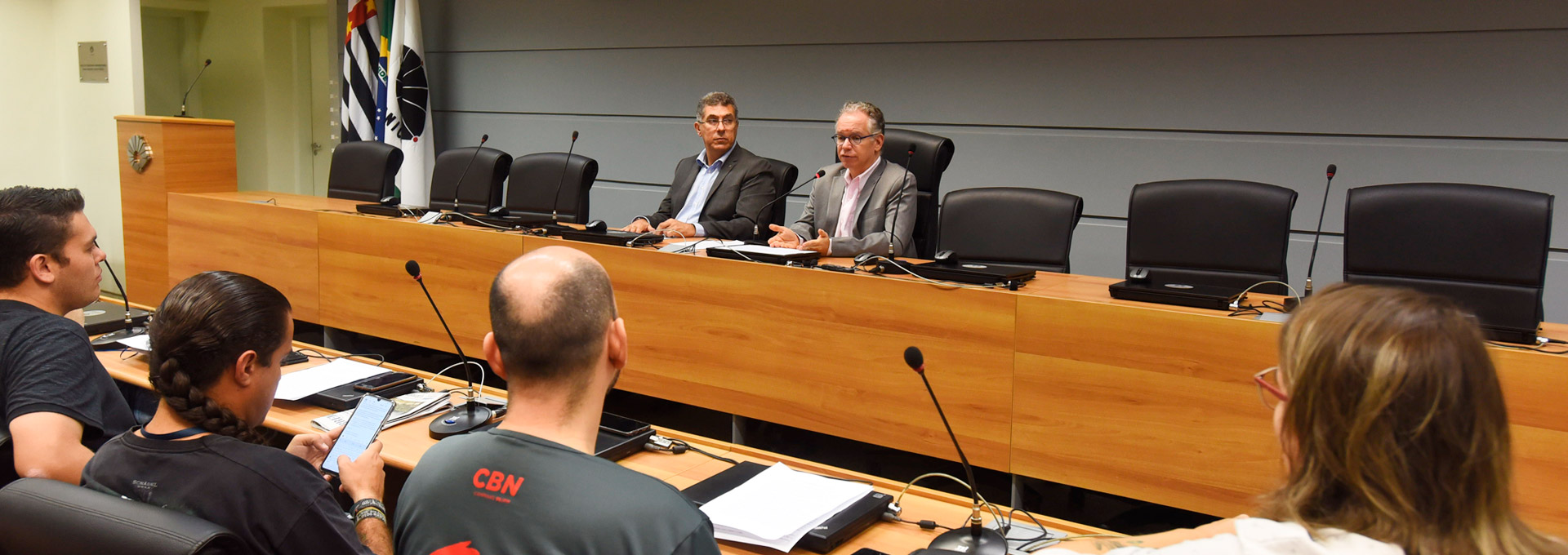 Unicamp aprova moção contra cortes no financiamento de pesquisa. Foto:Antonio Scarpinetti Audiodescrição: em amplo auditório, imagem em perspectiva e de corpo inteiro, dois homens sentados em poltronas, um ao lado do outro, e com olhar voltado para a esquerda da imagem. À frente deles, uma extensa mesa retangular de madeira onde ambos apoiam os braços. O homem à direita fala em microfone de mesa. Mais à frente deles, de costas na imagem, o público do auditório, com quatro pessoas sentadas em cadeiras dispostas uma ao lado da outra, formando uma fileira horizontal. Imagem 1 de 1.