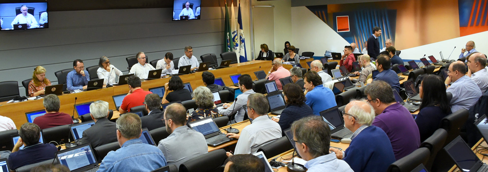 Unicamp convoca assembleia extraordinária em defesa da Ciência e Educação. Foto: Antoninho Perri Audiodescrição: no palco de um auditório, imagem panorâmica e em perspectiva, seis pessoas sentadas em cadeiras, uma ao lado da outra, com olhares voltados para a direita da imagem. A segunda pessoa à direita fala em microfone de mesa. A maioria delas apoia os braços sobre uma extensa mesa retangular de madeira posicionada à frente, sobre a qual há notebooks abertos e microfones de mesa. À frente deles, de costas na imagem, o público do auditório, cerca de trinta pessoas sentados em cadeiras. Imagem 1 de 1.