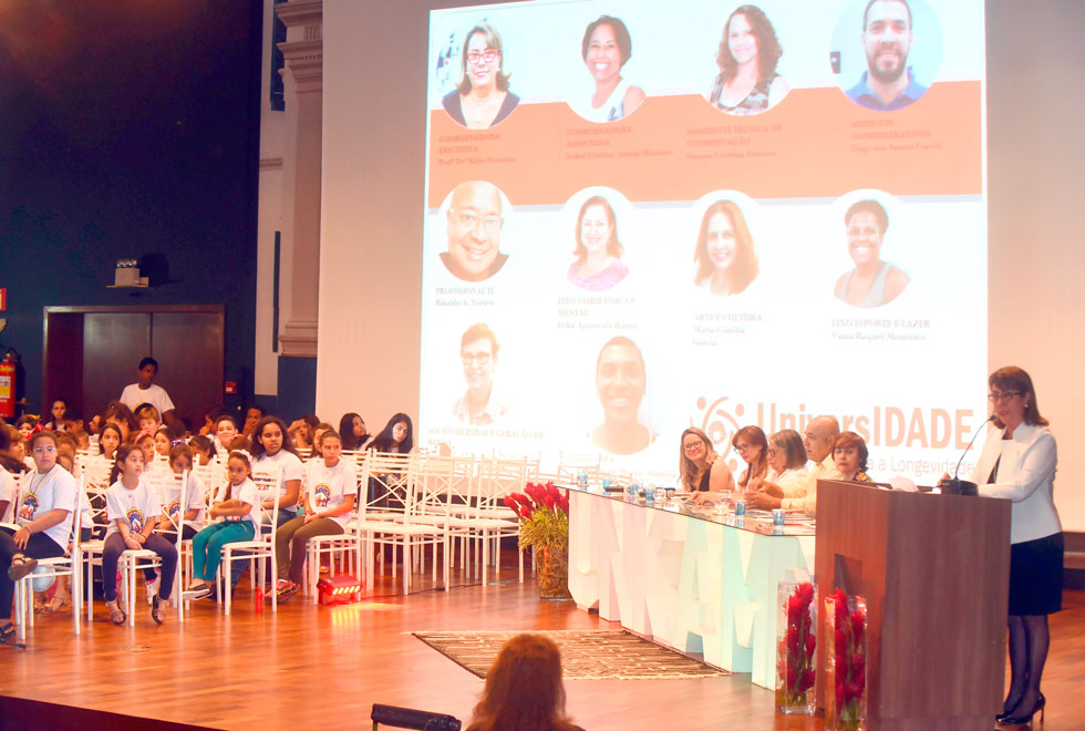 Cena mais aberta do palco em que vemos o telão iluminado com fotos da equipe do programa UniversIDADE