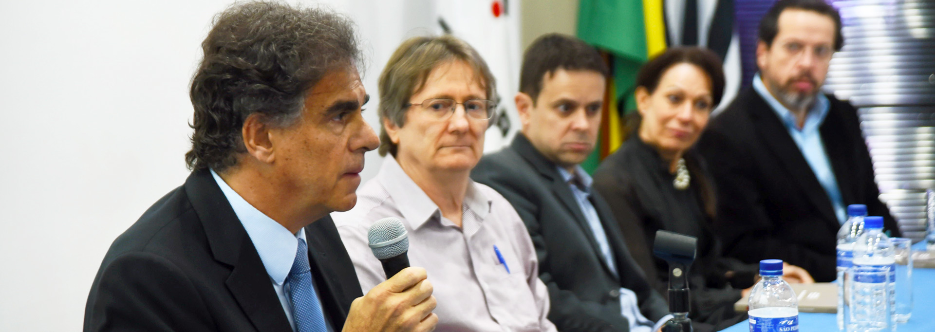Mesa de debate do seminário sobre “A realidade após a implementação da reforma trabalhista”, no Instituto de Economia. Foto: Antonio Scarpinetti Em auditório, imagem em perspectiva e de busto, cinco pessoas sentadas em cadeiras, uma ao lado da outra, e voltadas para a direita da imagem. A primeira pessoa à esquerda na imagem fala em microfone sem fio. À frente deles, uma extensa mesa retangular, onde alguns apoiam os braços. Sobre a mesa há garrafas pet pequenas de água e copos de vidro. Ao fundo da imagem, duas bandeiras de tecido hasteadas em pequenos mastros. A maioria das pessoas veste roupa social, como blazer e camisa de botão. Imagem 1 de 1.