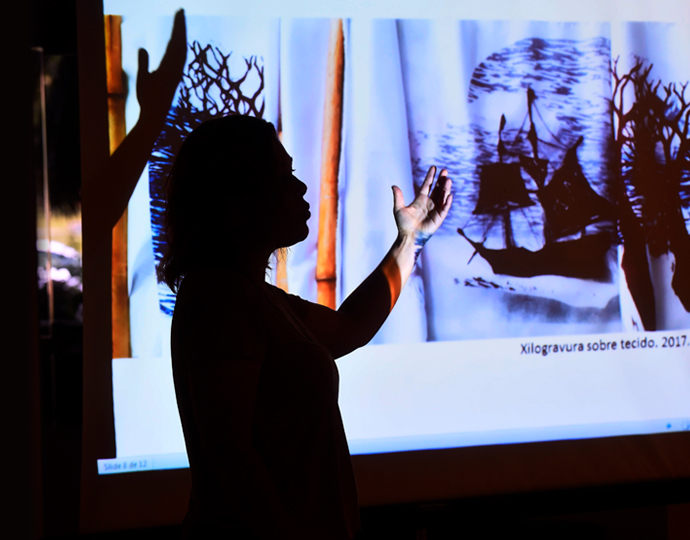 Audiodescrição: fotografia colorida mostra silhueta da mestranda Ana Paula Rodrigues de Melo apontando para uma tela de projetada em que aparece uma pintura sobre tecido com a temática de barcos. Ela está no meio de sua defesa de dissertação.