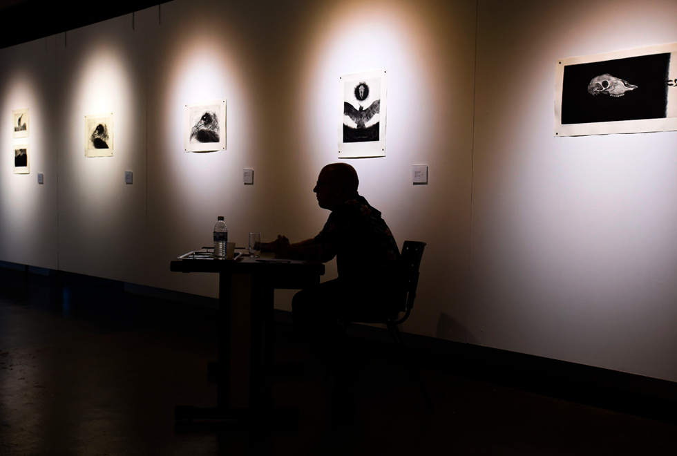 Artista André Berger, durante defesa de mestrado; ao fundo, parte de sua exposição. Audiodescrição: fotografia colorida mostra silhueta de André Berger sentado e olhando para a frente. A fotografia é escura e ao fundo, iluminadas, estão cinco de seus trabalhos, em preto e branco.
